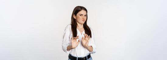Displeased woman showing stop, rejection gesture, waving hands to decline, refuse of something ugly, standing over white background photo