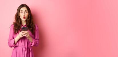 Image of surprised young woman gasping and saying wow, staring impressed at camera after reading news online on mobile phone, standing with smartphone against pink background photo