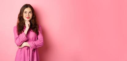 Stylish thoughtful woman with make up and spring dress, looking aside at logo and smiling pleased, thinking while standing against pink background photo