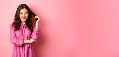 Beautiful brunette woman enjoying spring, wearing bright clothes and make up touching her curly hair and smiling confident at camera, standing against pink background photo
