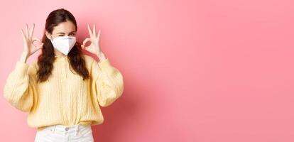 Covid-19, lockdown and pandemic concept. Young smiling girl assure you, winking and showing okay sign, alright or approve gesture, wearing medical respirator, standing over pink background photo