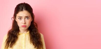Close up of cute and coy girl making sad pouting face to ask for something, wants pity, standing against pink background gloomy photo