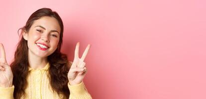 Close up of beautiful young woman showing peace v-sign and smiling happy at camera, wearing bright glam make up, standing against pink background photo