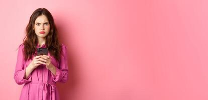 Angry woman frowning and looking confused after reading mobile phone news, standing puzzled and upset against pink background photo