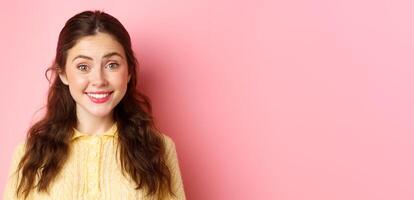 Close up of cute brunette lady smiles at camera, trying to make friendly and polite face, stands against pink background photo