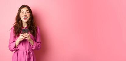 Beautiful brunette girl with amazed face, gasping and looking wondered, holding smartphone, see awesome news online, standing against pink background photo