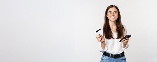 Smiling modern woman using credit card and mobile phone, paying, making purchase online, shopping on smartphone, standing over white background photo