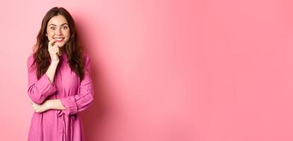Beautiful excited girl smiling, biting finger and looking with tempted face at camera, having interesting idea, looking at something intriguing, standing against pink background photo
