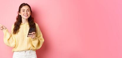 Beautiful modern girl holding smartphone and pointing finger aside, showing promotional text on copyspace while using mobile phone, standing over pink background photo