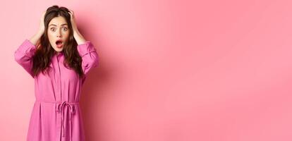 Shocked glamour girl gasping and looking startled, holding hands on head, standing in panic against pink background photo