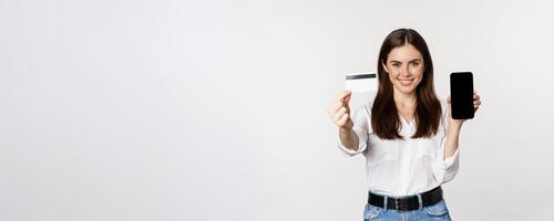 Portrait of female model showing credit card with smartphone screen, recommending application, standing over white background photo