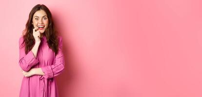 Beautiful excited girl smiling, biting finger and looking with tempted face at camera, having interesting idea, looking at something intriguing, standing against pink background photo
