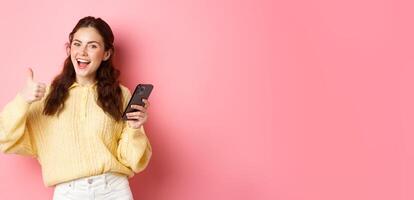 Portrait of attractive glamour girl showing thumbs up after using app on smartphone, online shopping application, standing against pink background, smiling satisfied photo