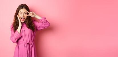 Positive brunette girl partying, looking happy, touching face and showing v-sign, standing excited against pink background photo