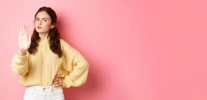 Angry and bossy young woman frowning, looking serious, showing block stop gesture, stretch out hand to say no, refuse something bad, forbid action, standing against pink background photo