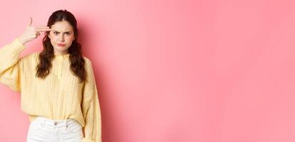 Annoyed and bothered girl frowning, showing finger gun next to her forehead, something annoying blowing her mind, standing against pink background photo