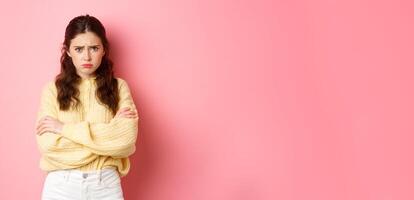 Cute silly girl feeling offended and sad, trying to comfort herself with self hug, frowning and looking at camera insulted, about to cry, standing against pink background photo