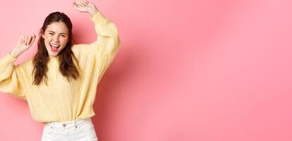 Image of beautiful caucasian woman having fun, dancing and enjoying party, raising hands up in relaxed, carefree pose, standing against pink background photo