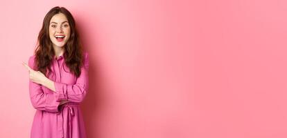 Stylish young modern woman look amazed, pointing finger left aside at copyspace, showing promo text, standing happy against pink background photo