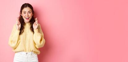 Lucky cute girl cross fingers and smiling, hope for dream come true, making wish, waiting exam results and looking hopeful at camera, standing over pink background photo
