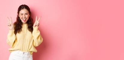 Cute and positive young woman winking, showing tongue, making v-sign gseture and smiling, standing upbeat against pink background photo