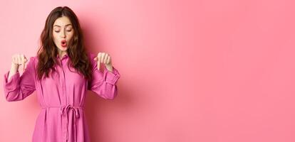 Beautiful female model in spring outfit, pointing and looking down, showing copyspace aside, standing against pink background photo