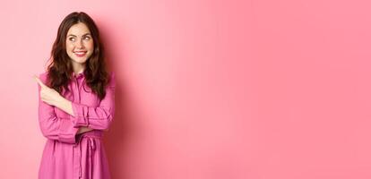 Beautiful glamour girl with make up, wearing spring outfit, pointing and looking aside at logo, showing banner on left copyspace, standing against pink background photo