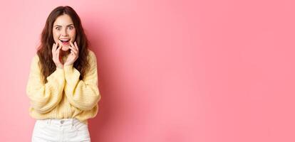 Omg really. Excited young woman hear gossip or super good news, staring amazed, smiling and holding hands on face, listen to interesting rumor, standing over pink background photo