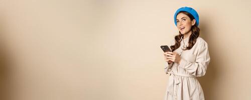 Modern girl using smartphone, smiling and posing happy with mobile phone, standing over beige background photo