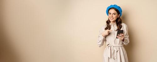Image of young stylish french woman in trench coat, standing with smartphone, using mobile phone app and smiling, posing against beige background photo