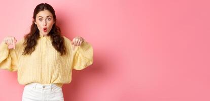Wow super cool. Excited brunette girl pointing fingers down, gasping wondered, showing direction way, indicating at bottom logo, standing over pink background photo