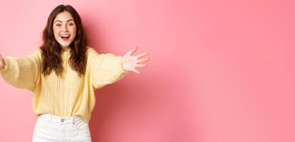 Portrait of beautiful friendly girl stretch out hands and smiling, reaching towards camera to give hug, making warm welcome, standing against pink background photo