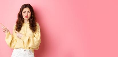 Sad and gloomy brunette girl complaining on bad promotional text, pointing fingers aside to left, showing banner with disappointed expression, standing over pink background photo