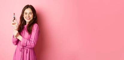 Coquettish smiling woman showing lipstick, applying make up for party event, standing in dress against pink background photo