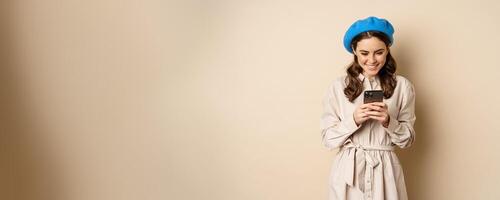 Modern girl using smartphone, smiling and posing happy with mobile phone, standing over beige background photo