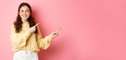 Look this way. Smiling beautiful female model pointing fingers right at copyspace aside, showing advertisement, looking at camera upbeat, standing against pink background photo
