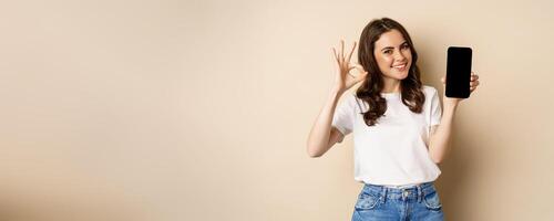 Smiling happy girl showing mobile phone screen, app on smartphone and okay, recommending online store, shopping application, beige background photo