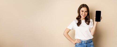 Online shopping and app concept. Young woman smiling and showing mobile phone screen, application interface, standing over beige background photo
