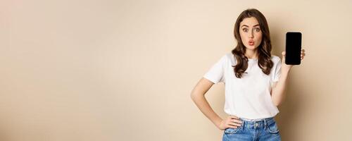 Online shopping and app concept. Young woman smiling and showing mobile phone screen, application interface, standing over beige background photo