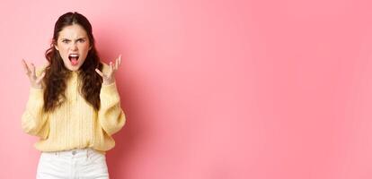 Angry and pissed-off girl screaming, shaking hands and grimacing furious, stare with outraged annoyed face at camera, standing against pink background photo