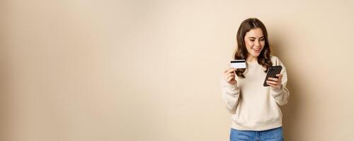Smiling girl using mobile app, smartphone shopping and credit card, standing over beige background, order smth photo