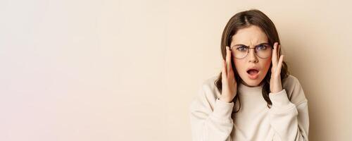 Close up portrait of woman in glasses looking confused and frustrated, cant understand smth strange, standing over beige background photo