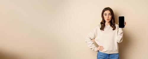 Stylish smiling woman showing smartphone screen, mobile app interface, standing over beige background photo