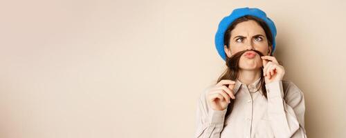 Funny beautiful girl fooling around, making moustache out of hair under lips, squinting and grimacing, standing happy against beige background photo