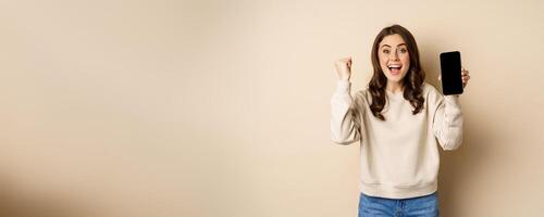 Enthusiastic woman cheering and showing cellphone screen, smartphone app interface, standing over beige background photo
