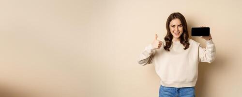 Smiling happy woman showing smartphone horizontal screen, thumbs up, recommending website, online store or app, standing over beige background photo