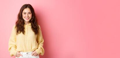 Portrait of good-looking young woman in sweater, holding hands in pockets, smiling and staring determined at camera, standing against pink background photo