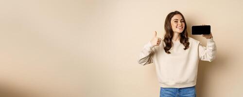 Smiling happy woman showing smartphone horizontal screen, thumbs up, recommending website, online store or app, standing over beige background photo