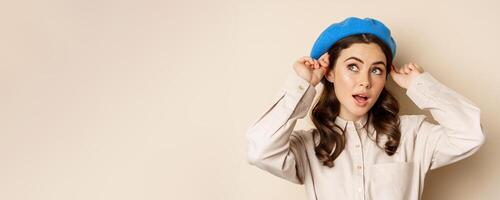 Stylish modern girl put on trendy hat on head and smiling, going out, posing against beige background photo