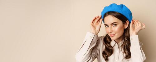 Stylish modern girl put on trendy hat on head and smiling, going out, posing against beige background photo